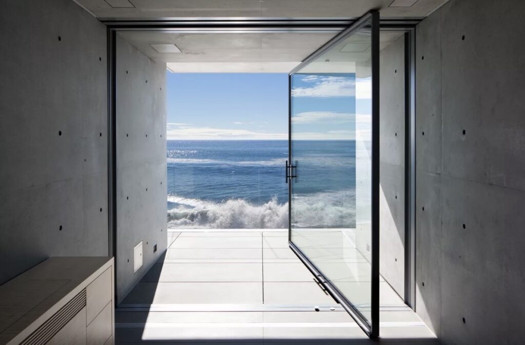 An open glass pivot door in a minimalist concrete room frames a breathtaking view of crashing ocean waves under a bright blue sky.